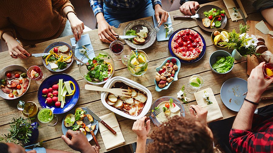 Gedeckter Holztisch mit vegetarischen Speisen, um den Menschen sitzen, die nur teilweise zu sehen sind